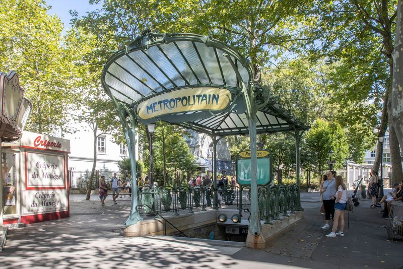The green glass-and-metal canopy of the Abbesses metro entrance beneath trees.