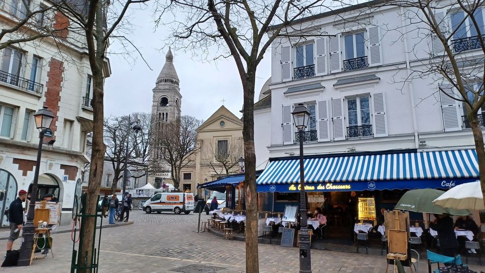 Café terraces and cobblestones on Place du Tertre, with Saint-Pierre church and the Sacré-Cœur tower beyond.