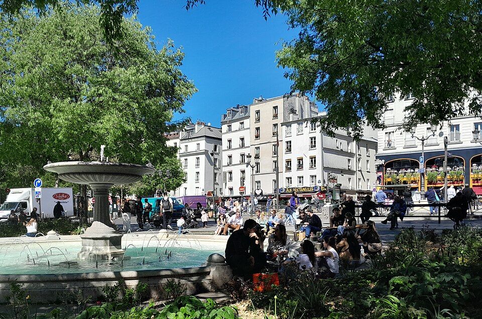 The fountain on Place Pigalle beneath spring trees, with people sitting nearby and apartment buildings beyond, photographed in April 2026.